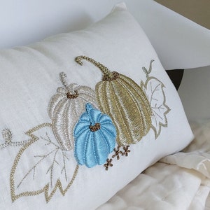 May include: A rectangular white pillow with an embroidered design of three pumpkins and leaves. The pumpkins are silver, gold, and blue. The leaves and stems are gold. The pillow is resting on a white surface.