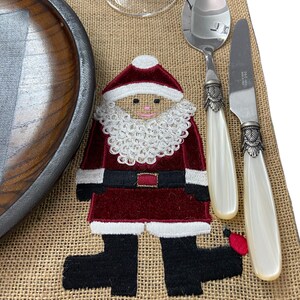 May include: A burlap placemat with an embroidered Santa Claus design. The Santa is wearing a red suit with white trim and a white beard. The placemat is set with a wooden plate, a glass, a spoon, and a knife.