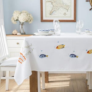 Può includere: Tovaglia bianca con ricamo a tema vita marina. Presenta pesci colorati e coralli. La tavola è apparecchiata per un pasto, con piatti, bicchieri e una mappa sulla parete sullo sfondo.