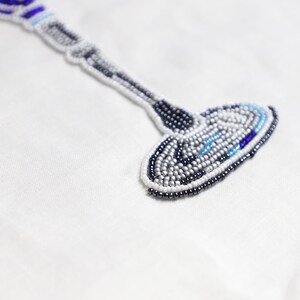 May include: A white fabric background with a beaded design of a wine glass. The glass is made of white, blue, and silver beads.