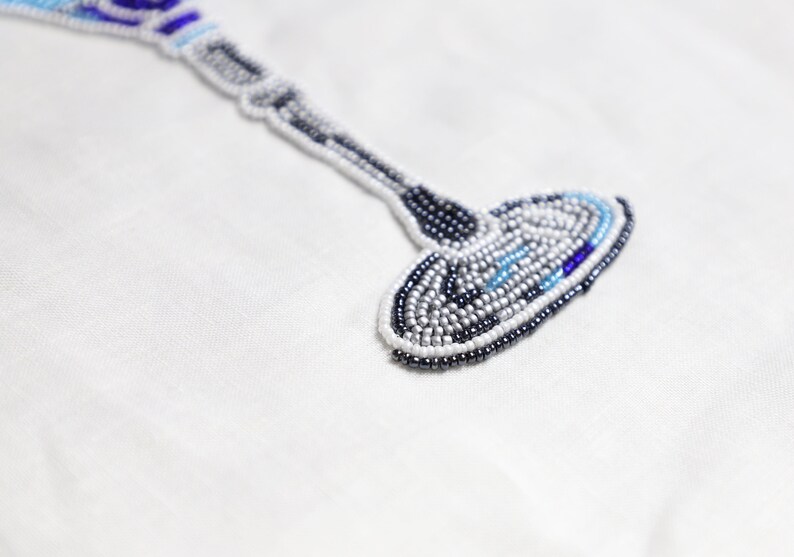 May include: A white fabric background with a beaded design of a wine glass. The glass is made of white, blue, and silver beads.