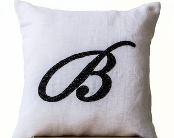 May include: Square white pillow with a large, black, cursive letter "B" embroidered on the front. The pillow has a textured appearance, suggesting a linen or similar fabric. The black embroidery provides a striking contrast against the white background.