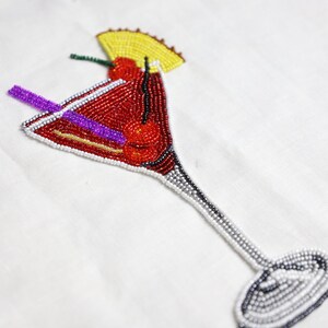 May include: A beaded cocktail glass with a red cherry and a purple straw. The glass is made of white and silver beads.
