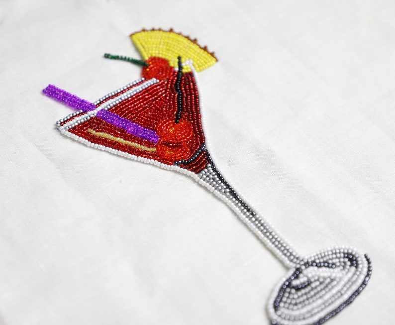 May include: A beaded cocktail glass with a red cherry and a purple straw. The glass is made of white and silver beads.