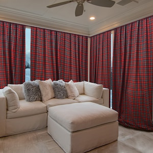 Puede incluir: Cortinas a cuadros rojos y negros enmarcan una ventana luminosa, complementadas con un sofá seccional blanco y una otomana. Cojines decorativos en gris y blanco añaden textura. Una pared de ladrillo y un taburete estampado completan el salón.