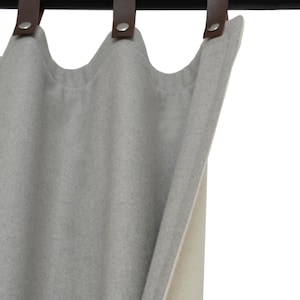 Puede incluir: Una cortina gris claro con superficie texturizada, colgada de una barra negra con correas de cuero marrón. La cortina tiene un borde superior ondulado y un reverso beige claro.