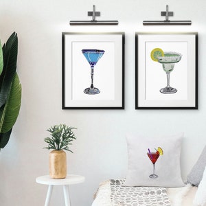 May include: Three framed illustrations of cocktails. The first is a blue martini, the second is a green margarita with a lime wedge, and the third is a red cosmopolitan with a lime wedge and a cherry.