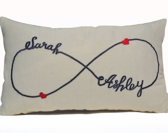 May include: Cream-coloured rectangular cushion embroidered with the names "Sarah" and "Ashley" in navy blue script, linked by an infinity symbol. Small red hearts accent the design.
