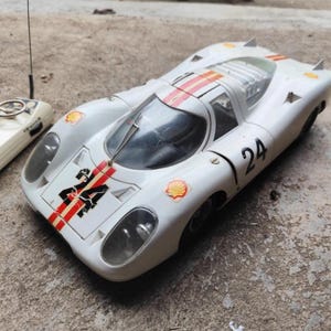 May include: A white vintage toy race car with the number 24 on the hood and side. The car has red and orange stripes and Shell logos. A white remote control is visible to the left.
