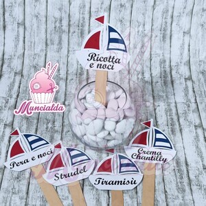 Può includere: Un set di cinque topper per torte a forma di barca a vela bianca e blu con diversi nomi di dessert scritti su di essi. I nomi sono Ricotta e noci, Pera e noci, Strudel, Tiramisù e Crema Chantilly. I topper sono su bastoncini di legno e sono disposti su una superficie di legno.