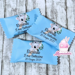 Può includere: Tre incarti per caramelle azzurri con pois bianchi, ciascuno con un'illustrazione di koala e il testo Riccardo Il mio Battesimo 20 Giugno 2021. Un logo a forma di cupcake rosa con la scritta Muncialda è posizionato vicino agli incarti.