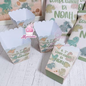Può includere: Un set di scatole di carta bianca per snack con un tema di dinosauri e illustrazioni verdi e beige. Ogni scatola riporta la scritta IL 3° COMPLEANNO DI NOAH, accompagnata da vari dinosauri a cartoni animati, tra cui un triceratopo e uno pterodattilo.