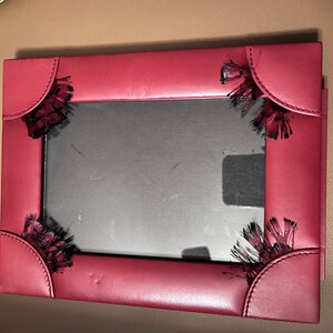 May include: A red leather picture frame with black fringe detailing around the edges. The frame is slightly damaged, with some of the fringe missing.