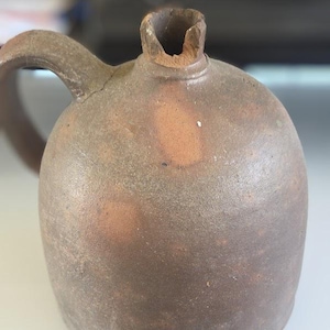 May include: A brown, antique-style stoneware jug with a rounded body and a handle. The jug has a wide opening and a textured surface, suggesting it is handmade. The color is a mix of brown and tan.