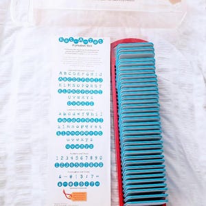 May include: A Sizzix Rat-A-Tat Alphabet Set with a red and blue design. The set includes alphabet letters, numbers, and symbols. The letters are in a blue font. The set is in a clear plastic case.