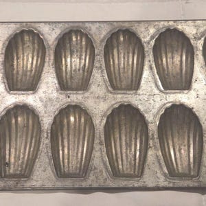 May include: A rectangular, vintage-style madeleine pan with 12 shell-shaped molds. The pan is made of a dull, silver-colored metal and sits on a white surface. Each mold has a ribbed interior.