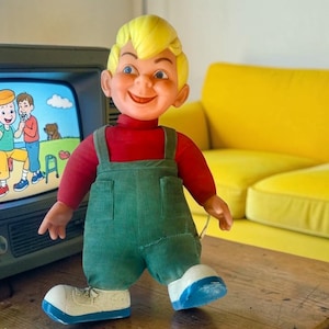 May include: A vintage doll with blonde hair, red shirt, and green overalls stands in front of a retro television. The TV screen displays a cartoon scene. A yellow sofa is in the background.