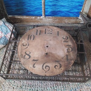 May include: A vintage clock face with black numerals on a distressed, aged brown background. The clock face sits inside a rectangular metal wire basket. The numbers are in a classic serif font. The clock face is a decorative item.
