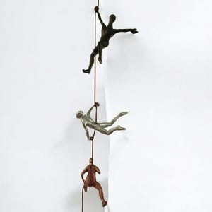 May include: Three bronze-toned climbing figures ascend a rope against a white wall. The figures are in various climbing poses, with outstretched arms and legs. The rope is a dark brown colour.