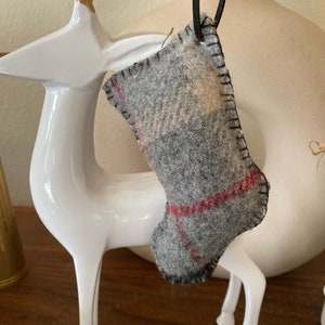 Pode incluir: Uma pequena meia de Natal cinza de feltro com detalhes xadrez vermelhos e costuras pretas. A meia é suspensa por um cordão preto. O fundo inclui uma estatueta de veado branca e um vaso creme.