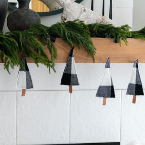Puede incluir: Una guirnalda festiva de árboles de Navidad de tela con patrones de cuadros en blanco y negro. Cada árbol está sujeto a una cuerda con una pequeña base de madera. La guirnalda se exhibe contra una pared de azulejos blancos, con vegetación y objetos decorativos en el fondo.