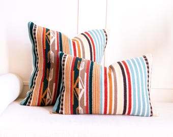 May include: Two rectangular throw pillows featuring a woven pattern of vertical stripes in teal, orange, red, brown, cream, and light blue. The design includes geometric arrow-like motifs integrated into the multi-colored striped fabric.