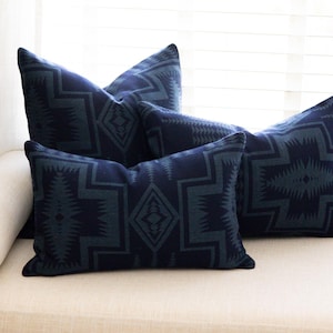 May include: Four navy blue throw pillows with a geometric pattern in a lighter shade of blue. The pillows are arranged on a cream-colored sofa near a window, creating a cozy and stylish interior design.