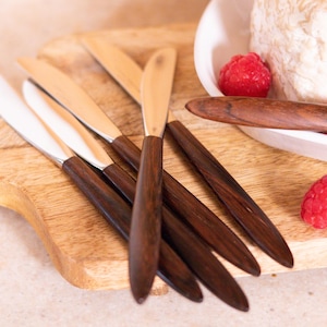 Könnte beinhalten: Ein Set aus sechs Buttermessern mit Holzgriffen und silbernen Klingen. Die Messer sind auf einem Holzbrett mit einem weißen Teller und roten Himbeeren im Hintergrund angeordnet.
