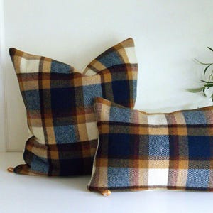 Op de afbeelding: Twee geruite kussens in blauw, bruin en crème. Het grotere kussen staat rechtop, het kleinere horizontaal. Beide hebben een klassiek, rustiek ontwerp. Ideaal voor een bank of bed.
