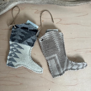 Puede incluir: Dos pequeños adornos de botas de vaquero. Uno es gris y negro con un patrón geométrico, el otro es marrón con un diseño similar. Ambos tienen un lazo para colgar y están sobre una superficie de madera clara.
