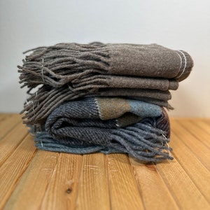 May include: A pile of folded wool scarves in shades of grey and brown. Each scarf has a fringed edge and is neatly folded. The scarves are stacked on a wooden surface.
