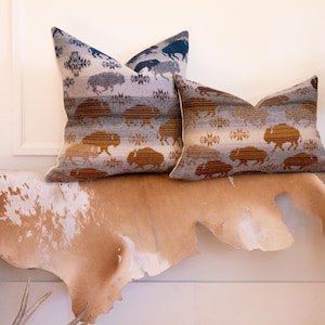 May include: Two decorative cushions featuring a bison pattern in brown, grey, and blue hues. They are placed on a rustic wooden bench with a cowhide throw. The cushions have a geometric design.