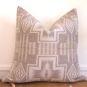 Puede incluir: Un cojín decorativo cuadrado con un patrón geométrico en tonos grises y blanquecinos. El cojín presenta un diseño repetitivo de diamantes, rectángulos y formas estilizadas de árboles.