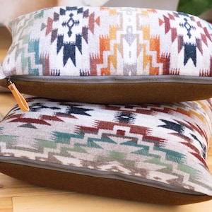 Puede incluir: Dos almohadas decorativas con un patrón del suroeste. Las almohadas están hechas de una tela suave y tejida y tienen una cremallera. Las almohadas están apiladas una encima de la otra, con la almohada superior que presenta un patrón más colorido.