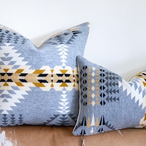 May include: Two decorative cushions with a geometric pattern in shades of grey, white, black, and gold. The cushions have a soft, textured appearance and are placed on a brown surface.