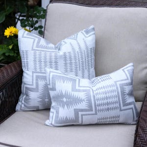 May include: Two decorative throw pillows with a white background and a gray geometric pattern. The pillows are rectangular and square, resting on a beige cushion. The pillows have a tribal design.