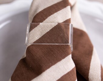Op de afbeelding: Een rechthoekige servetring van helder acryl die een gevouwen stoffen servet vasthoudt met een patroon van dikke, diagonale bruine en beige strepen. De servetring heeft een gladde, transparante afwerking en een eenvoudig geometrisch ontwerp.