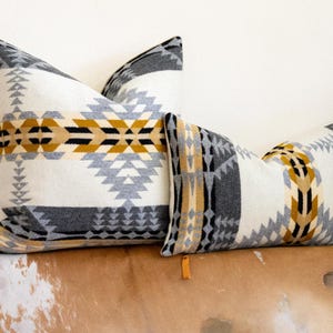 May include: Two decorative throw pillows with a geometric pattern in shades of white, grey, and yellow. The pillows are on a brown and white cowhide rug.