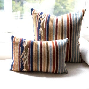 Puede incluir: Dos cojines, uno rectangular y otro cuadrado, con un patrón tejido de rayas verticales en tonos azul marino, naranja óxido, beige, crema y azul claro. El diseño incluye un motivo geométrico de flechas repetido en el lado izquierdo.