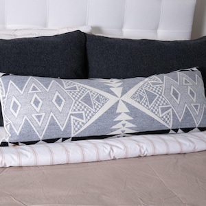 May include: A long, rectangular decorative pillow with a geometric pattern in shades of gray and white, accented with black trim. The pillow is placed on a bed with dark gray pillows and a striped comforter.