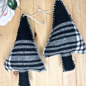 Pode incluir: Duas decorações de Natal artesanais em forma de árvore. Cada árvore é feita de tecido xadrez preto e branco com um tronco preto liso. Costuras brancas delineiam a forma da árvore. Uma alça de couro é presa para pendurar. Em uma superfície de madeira.