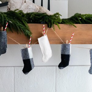 Puede incluir: Una colección de medias navideñas en miniatura en varios patrones y colores, incluyendo cuadros blancos y negros, blanco liso y gris. Las medias están suspendidas de una viga de madera con hilo y pinzas, y decoradas con pajitas a rayas rojas y blancas.
