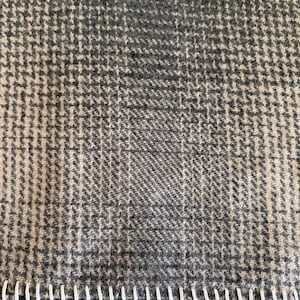 May include: Close-up of a gray and cream-colored fabric with a houndstooth pattern. The fabric has a white stitched edge along the bottom. The material appears to be a woven textile, possibly wool or a wool blend.
