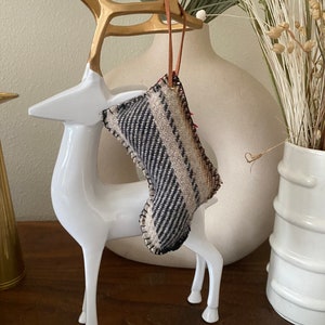 Pode incluir: Um enfeite decorativo de meia de Natal com uma alça de couro marrom pendurada em uma estatueta de veado branca com chifres dourados. A meia é feita de tecido tecido em tons de preto, bege e creme.