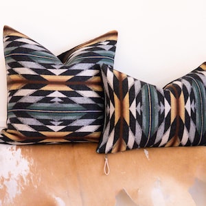 May include: Two decorative throw pillows with a geometric pattern in shades of black, white, green, and brown. The pillows are on a light brown surface.