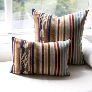 Op de afbeelding: Twee sierkussens met een geweven patroon van verticale strepen in marineblauw, roestoranje, beige en crème. Het ontwerp bevat geometrische pijlmotieven aan de linkerkant van elk kussen, afgewerkt met een zachte, gestructureerde stof.