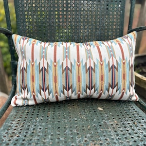 Op de afbeelding: Een rechthoekig kussen met een geometrisch patroon van blauwe, bruine, rode en gele pijlen op een witte achtergrond.