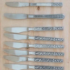 May include: Seven silver-coloured knives with ornate handles. The handles display floral and scrollwork patterns. The blades are smooth and curved. The knives are arranged on a light brown wooden surface, ideal for a dining setting.