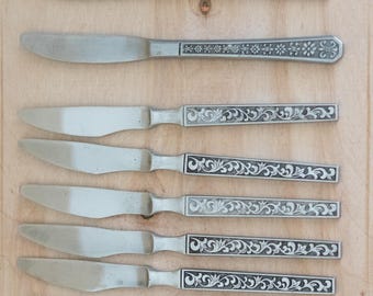 Op de afbeelding: Een verzameling van zeven zilverkleurige messen met decoratieve handvatten. De handvatten hebben ingewikkelde bloemen- en krulwerkontwerpen. De messen liggen op een lichtbruin houten oppervlak.
