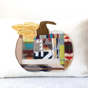 May include: A white cushion featuring a patchwork pumpkin design. The pumpkin has a brown stem and yellow leaf, with a variety of fabric patterns in shades of blue, green, red, and grey. The cushion is a decorative item.